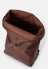 Mochila de cuero marrón con un diseño de tapa enrollable, que cuenta con un anillo en D de metal en tonalidad dorada y un acabado suave y texturizado en el exterior.
