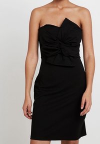 Robe noire sans bretelles avec un col asymétrique et un grand nœud central portée par une personne se tenant devant un fond uni.