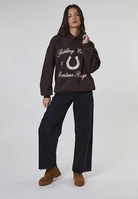 Brązowa bluza z kapturem z kremowym nadrukiem tekstu i wzorem podkowy, zestawiona z czarnymi spodniami o szerokich nogawkach oraz brązowymi sznurowanymi butami.