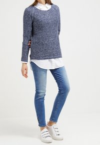 Personne portant un pull en maille bleu par-dessus une chemise blanche boutonnée, un jean skinny bleu clair avec des fermetures éclair aux chevilles, et des baskets blanches à scratch.