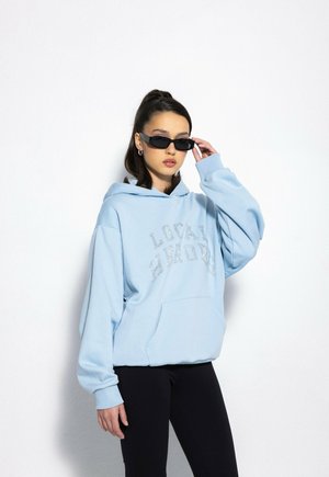 Jasnoniebieska bluza z kapturem z przednią kieszenią i oversize'owym krojem, z napisem w stylu kryształowym. W zestawie z czarnymi legginsami i okularami przeciwsłonecznymi.