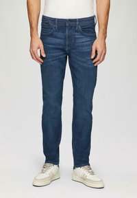 Dark blue denim jeans with a straight fit, five pockets, and slight fading. Paired with light-colored sneakers featuring beige and gray accents.