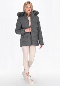 Giubbotto piumino grigio con cappuccio in pelliccia sintetica removibile, zip frontale, tasche laterali e design trapuntato. Abbinato a pantaloni attillati beige e sneakers.
