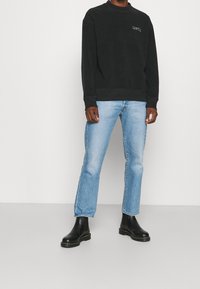 Uomo che indossa una felpa nera, jeans azzurri chiari e stivaletti in pelle nera, in piedi contro uno sfondo bianco semplice.