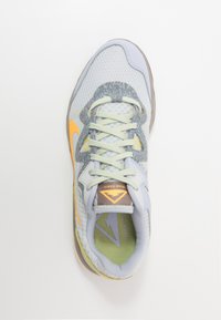 Scarpa sportiva grigio chiaro con tomaia in rete, dettagli in verde pastello, suola grigia texture e logo swoosh arancione. Presenta lacci piatti e colletto imbottito.
