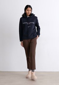 Marineblauwe hoodie met het "TOMMY HILFIGER" logo, bruine broek en beige schoenen. Model staat tegen een neutrale achtergrond.