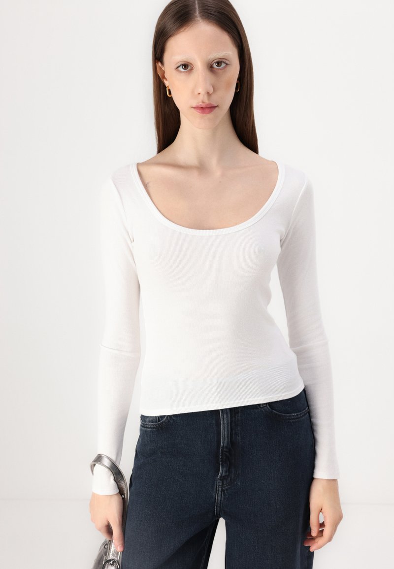 Weißes Langarmoberteil mit Rundhalsausschnitt und gerippter Textur, kombiniert mit dunklen Jeans. Minimales Design und figurnahe Silhouette.