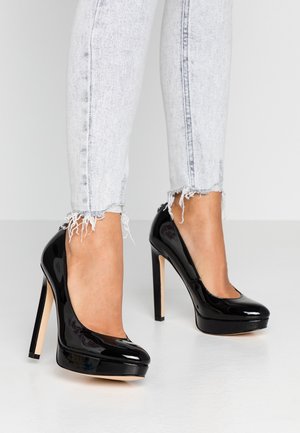 Legs wearing light gray frayed hem jeans and shiny black high-heeled platform pumps on a white floor and background.