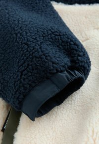 Tmavomodrý fleece materiál s textúrovaným povrchom, s hladkým elastickým manžetom. Na spodnej strane kontrastuje s krémovo farbeným plyšovým materiálom.