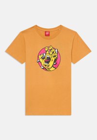 Orange bomullströja med ett grafiskt tryck av en gul hand och en öppen mun, omgiven av en rosa cirkel. Rund hals och korta ärmar.