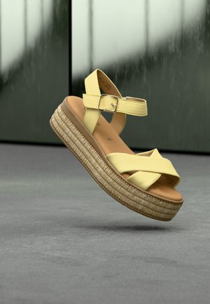 Sandale galbene din piele, cu barete încrucișate, cataramă la gleznă și talpă platformă împletită în stil espadrile. Plutind deasupra unei suprafețe gri.