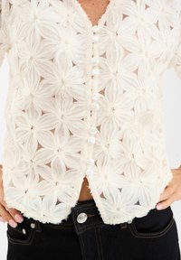 Balta ziedu mežģīņu blūze ar pērļu pogām, uzvilkta uz melnajām džinsām, rokas atpūšas uz gurniem, nagi nokrāsoti gaiši rozā.