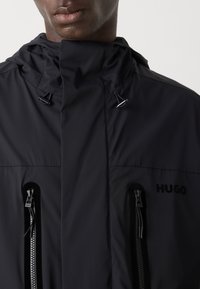Black lightweight jacket with a high collar, two zippered pockets, and a subtle logo. Features drawstrings for adjustable fit.