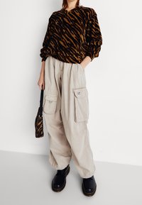 Brązowa i czarna bluza w panterkę w połączeniu z beżowymi spodniami typu cargo z bocznymi kieszeniami, noszona z czarnymi butami i wzorzystą torbą na ramię.