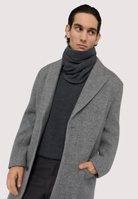 Šedý vlněný kabát v herringbone vzoru s širokým klopem, nosící se přes tmavě šedý svetr, doplněný pleteným, smyčkovaným šedým šátkem. Detail knoflíků na kabátu.