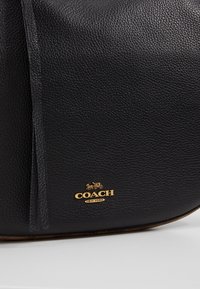 Gros plan sur un sac Coach en cuir noir texturé avec un petit logo doré et un emblème de calèche tirée par des chevaux près du bord inférieur.
