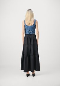 Denim prirezana majica s prilagodljivo zaponko, v kombinaciji z dolgo črno obleko s plasteh, ki se ponaša z gladko teksturo in razširjeno silhueto.