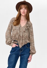 Kobieta w brązowym kapeluszu z szerokim rondem, bluzce w panterkę z falbankami, wpuszczonej w niebieskie dżinsy z wysokim stanem i beżowym paskiem, z ręką w kieszeni.