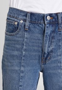 Vaqueros de mezclilla azul con cintura alta, con cierre de botones, cinco bolsillos y detalles de costura en color naranja. Textura suave con un diseño clásico.