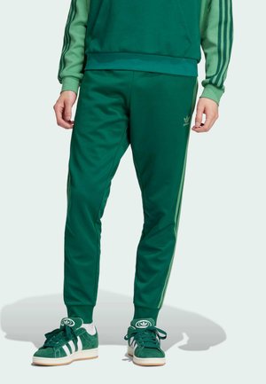 Pantaloni tuta verdi con due strisce verticali verdi, realizzati in tessuto morbido. Abbinati a sneaker verdi e bianche, con lacci e dettagli del logo.