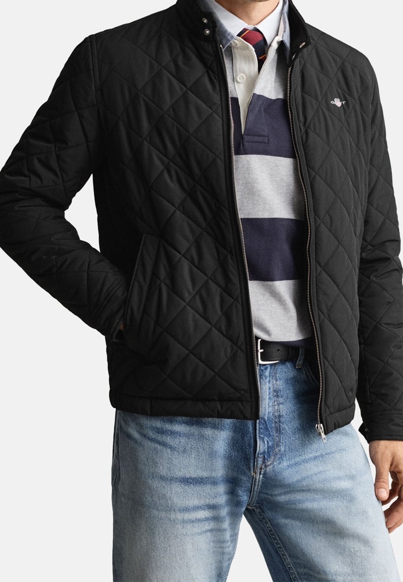Homme portant une veste matelassée noire ouverte sur un maillot de rugby rayé et un jean bleu clair, main dans la poche de la veste.