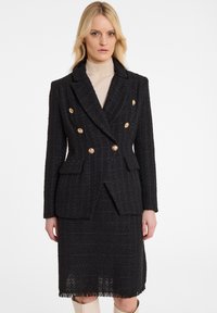 Zwarte tweed blazer met een dubbelgeknopte ontwerp, gouden knopen, zakken en textuurstof. Draagt over een lichtgekleurde top.