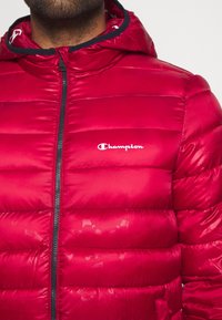 Rød pufferjakke med lynlås foran, hætte og Champion-logo på brystet. Har en kviltet design og en glat, blank overflade.