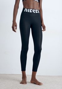 Leggings noirs en tissu lisse et extensible avec une taille élastique arborant le logo blanc "DIESEL". Design ajusté, longueur cheville.