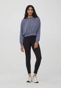 Lekki, krótki szary sweatshirt z długimi rękawami, elastycznym paskiem w talii, w połączeniu z czarnymi legginsami i białymi butami sportowymi.