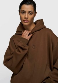 Mujer con sudadera marrón de gran tamaño sosteniendo el cuello con una mano, mirando al frente contra un fondo claro y liso.