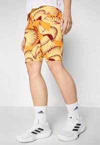 Oranžno raztegljive kratke hlače z vzorcem ptic, prilegajočega kroja, in roza Adidasovim logotipom. Nosi jih z belimi športnimi čevlji in nogavicami.