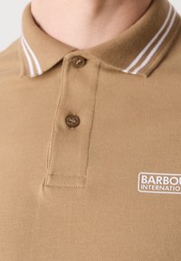 Polo couleur beige avec deux boutons marron et col à rayures blanches, arborant le logo Barbour International sur la poitrine.