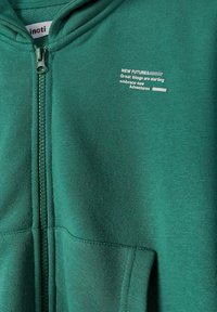 Sweat à capuche vert zippé avec poches avant et texte blanc sur la poitrine indiquant "NEW FUTURES De grandes choses commencent, embrassez de nouvelles aventures."