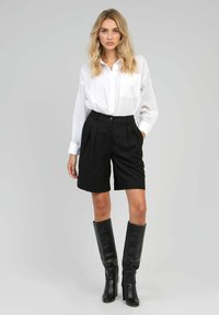 Chemise blanche à boutons, avec une poche poitrine, associée à des shorts noirs à rayures verticales et des bottes en cuir noires jusqu'aux genoux. Tissu lisse, coupe ajustée.