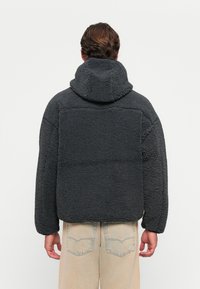 Dark gray fleece jacket with a hood, featuring a textured surface and contrasting stitching, worn over light beige pants, viewed from the back.