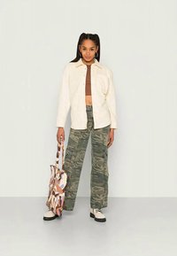 Camisa oversized bege, top curto castanho, calças cargo de camuflagem, bolsa tote creme com padrões abstratos e sapatos pretos com atacadores.