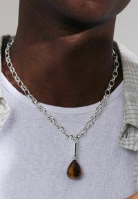 Silver chain necklace with large oval links, featuring a brown teardrop-shaped tiger's eye pendant. Worn over a textured grey shirt.