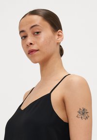 Svart spaghetti-top med en V-formad halslinje; mjukt, lätt tyg; har en blommig tatuering på modellens överarm.