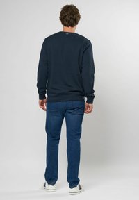 Marineblaues Sweatshirt mit langen Ärmeln, Rundhalsausschnitt, gerippten Bündchen und Saum; kombiniert mit dunkelblauen Jeans und weißen Sneakers mit Farbakzenten.