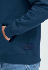 Personne portant un sweat à capuche bleu foncé avec une main dans la poche avant et un jean en denim bleu clair visible en bas.