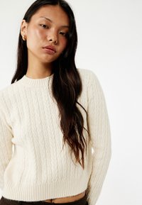 Suéter de punto trenzado en color crema con cuello redondo y puños acanalados, que presenta una textura suave y un ajuste ligeramente cropped.