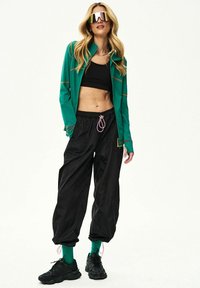 Žena oblečená v nadrozmerných čiernych nohaviciach, čiernom krátkom topu, zelenej bunde, zelených ponožkách, čiernych teniskách a veľkých reflexných slnečných okuliaroch stojaca na bielom pozadí.