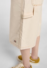 Pantaloncini cargo beige con cuciture arancioni, tasche laterali e una piccola etichetta vicino all'orlo. Indossati con sneakers in suede gialle.