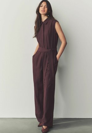 Mörkröd ärmlös jumpsuit med krage, sidofickor, tygbälte och en slät yta. Förstärkt med smala vertikala ränder.