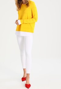 Maglione a righe gialle sopra una camicia bianca, abbinato a jeans bianchi e ballerine rosse a punta. Tessuto liscio e design casual.
