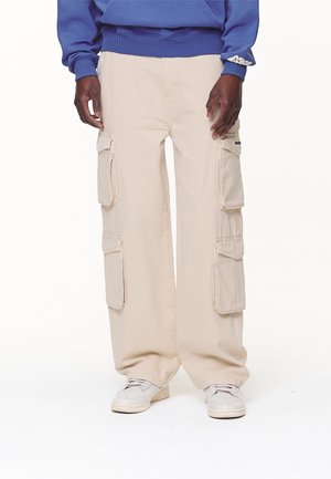 Person iført beige cargobukser med flere lommer, hvide sneakers og en blå sweatshirt med synligt manchetlogo.