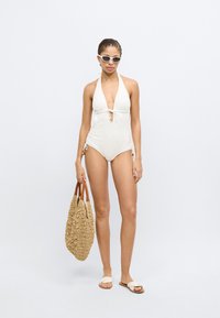 Λευκό ολόσωμο μαγιό με βαθύ V στο λαιμό και δέσιμο, υφασμένο σχέδιο, συνοδευόμενο από πλεκτή τσάντα ώμου και φλατ σανδάλια.