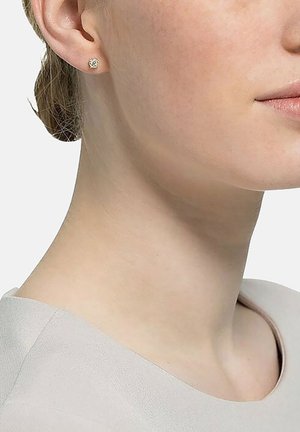 Goldene Steckerohrringe mit floralem Design, das eine strukturierte Oberfläche aufweist. Getragen an einem hellhäutigen Ohr, das die Details des Ohrrings hervorhebt.