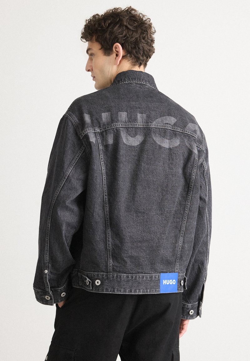 Mørk denimjakke med en afslappet pasform, der har et fremtrædende "HUGO" print på ryggen og et blåt mærke nederst.