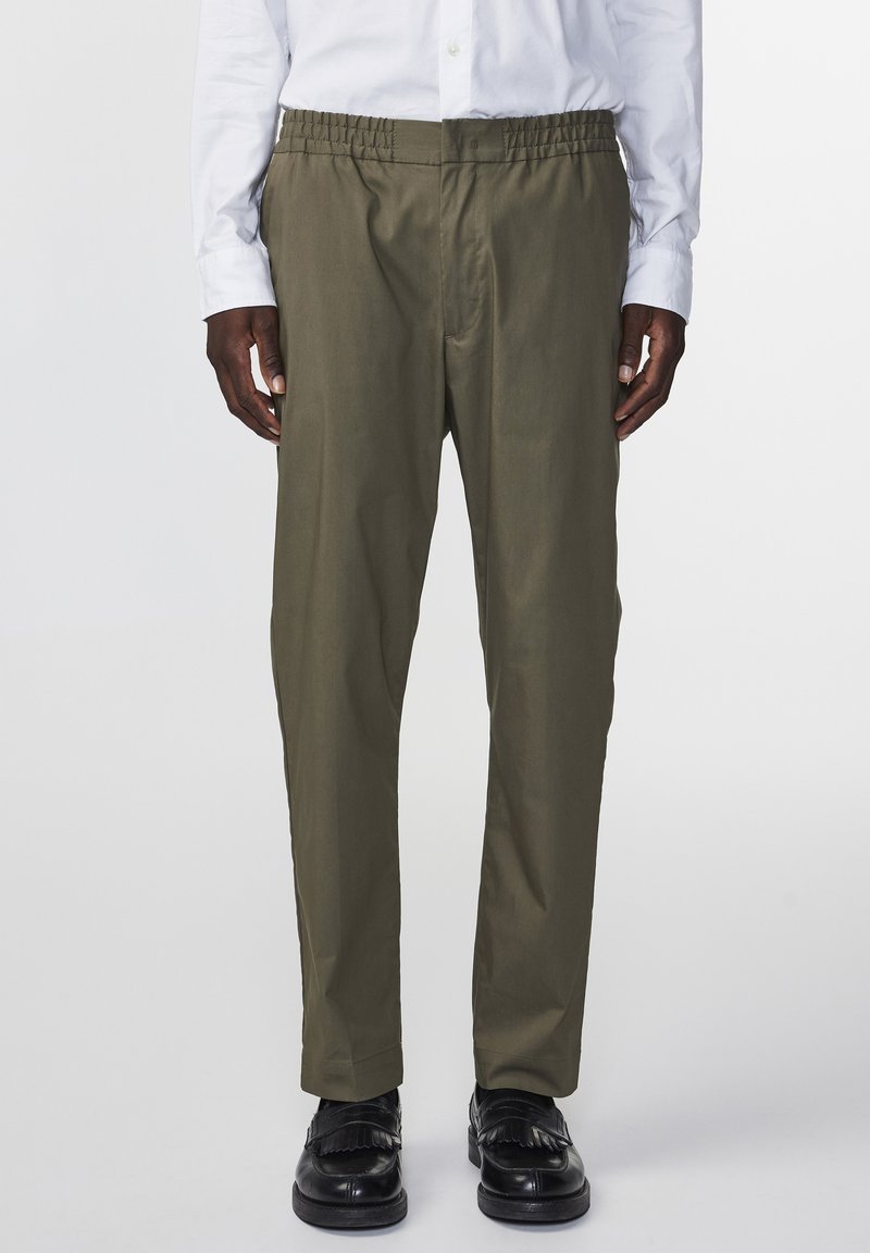 Pantalon vert olive avec taille élastique, coupe droite et tissu léger, associé à des chaussures noires avec des détails à franges.